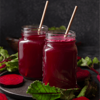 Foto de dois potes de vidro com suco de beterraba com canudo em um prato preto em cima de uma mesa preta com fatias e folhas de beterraba espalhadas