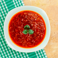 Foto com a visão superior de uma sopa de beterrraba em um prato fundo branco em uma mesa de madeira com uma toalha quadriculada verde